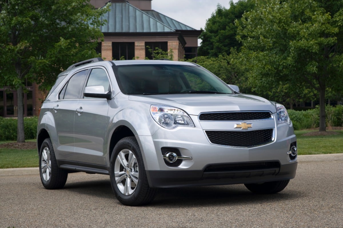 Image for: What are the Chevy Equinox Years to avoid? The image shows a silver Chevy Equinox in front of two green trees and a house