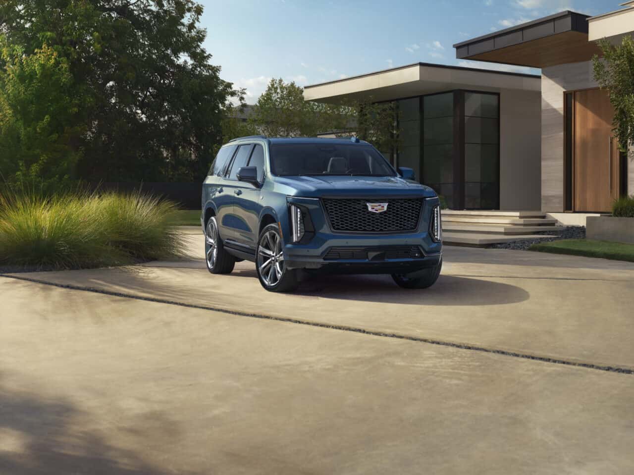 photo of Front ¾ view of the 2025 Cadillac Escalade Sport Platinum in Deep Sea Metallic with a house and greenery in the background.