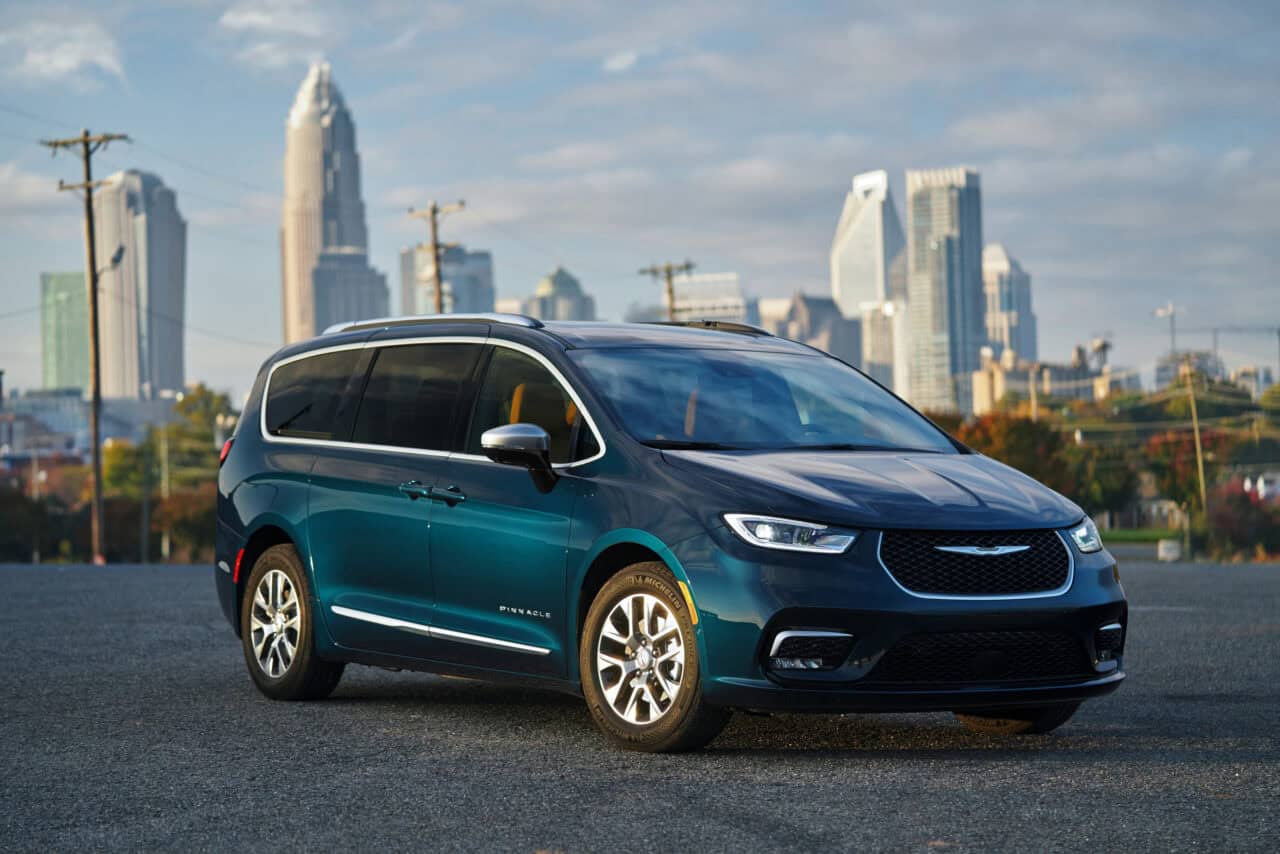 Photo of a 2025 chrysler pacifica hybrid sitting in front of a city landscape. It looks like the background might be Charlotte NC. 