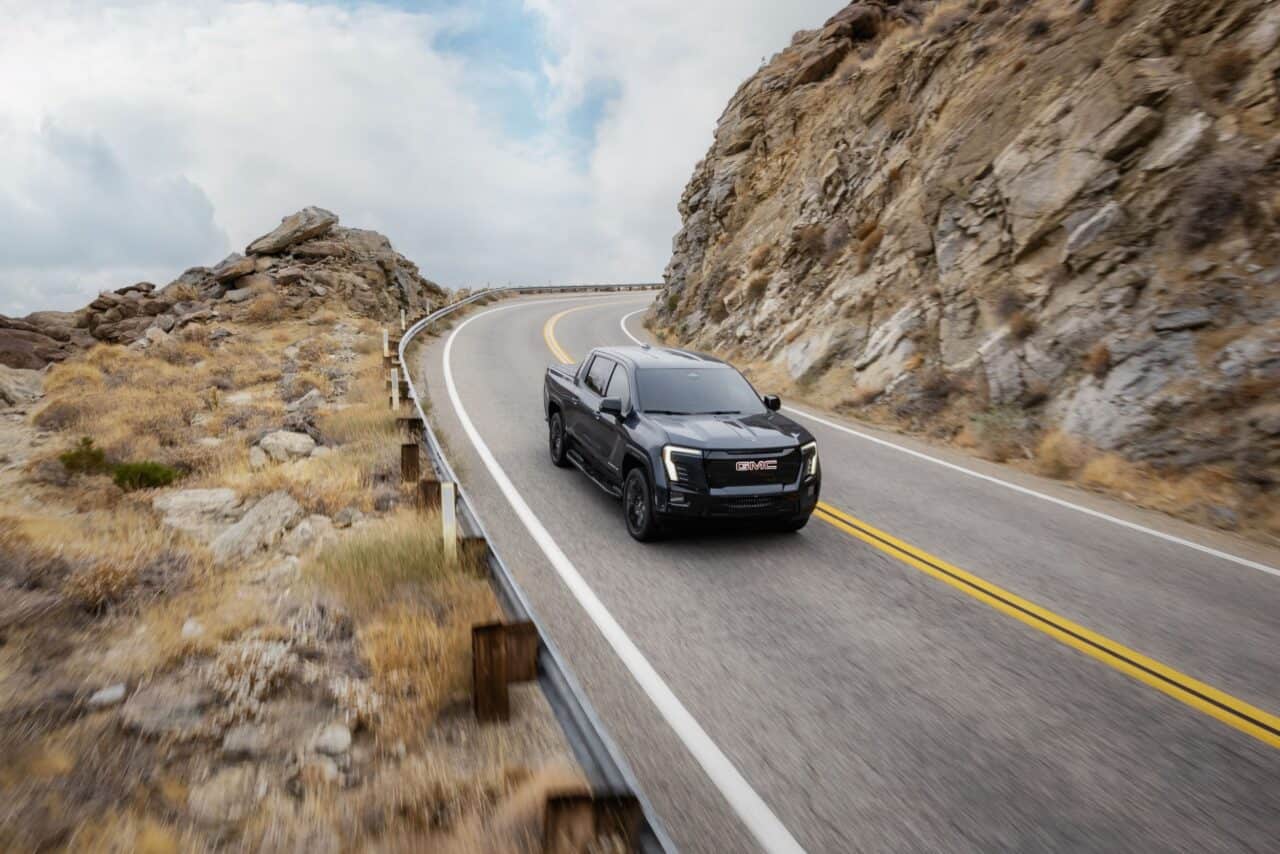 The 2026 Sierra Ev Elevation: Surprisingly Down To Earth 3 Front 3/4 shot of the 2026 GMC Sierra EV Elevation in Deep Ocean Metallic driving on a mountain road.